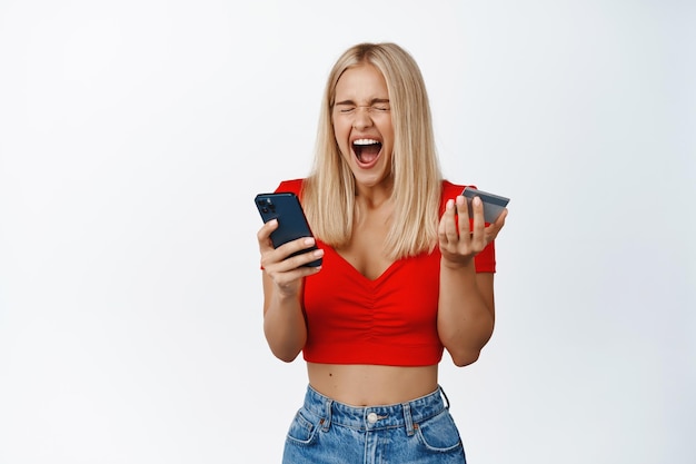 Aufgeregtes Glamour-Girl schreit, während es Smartphone und Kreditkarte in der Hand hält, erstaunt über den Online-Verkauf in der Shopping-App, die über weißem Hintergrund steht