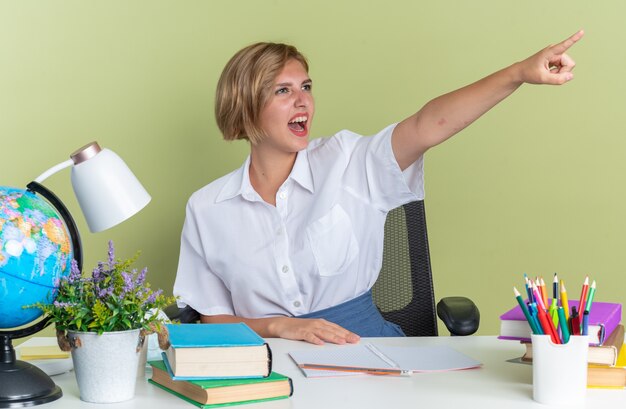 Aufgeregtes junges blondes Studentenmädchen, das am Schreibtisch mit Schulwerkzeugen sitzt und die Hand auf dem Schreibtisch hält und auf die Seite zeigt, die auf olivgrüner Wand isoliert ist?