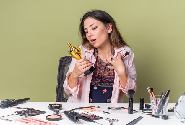 Aufgeregtes junges brünettes Mädchen, das am Tisch mit Make-up-Tools sitzt und Make-up-Pinsel hält und auf den Gewinnerpokal schaut