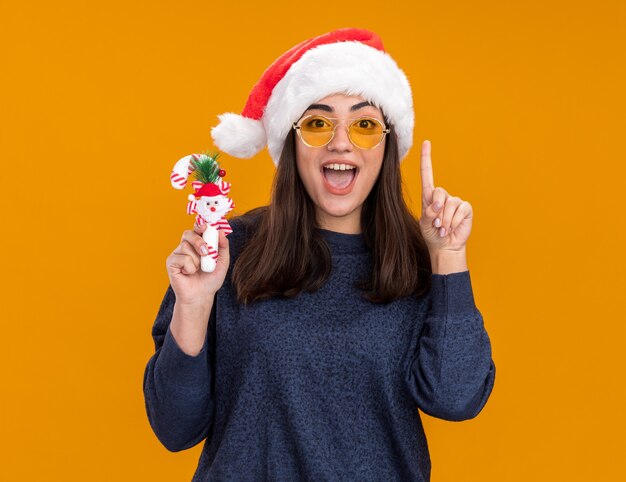 Aufgeregtes junges kaukasisches Mädchen in Sonnenbrille mit Weihnachtsmütze hält Zuckerstange und zeigt isoliert auf orangefarbener Wand mit Kopierraum