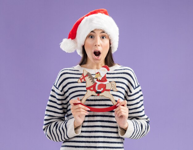 Aufgeregtes junges kaukasisches Mädchen mit Weihnachtsmütze, das den Weihnachtsmann auf Schaukelpferddekoration hält, isoliert auf lila Wand mit Kopierraum