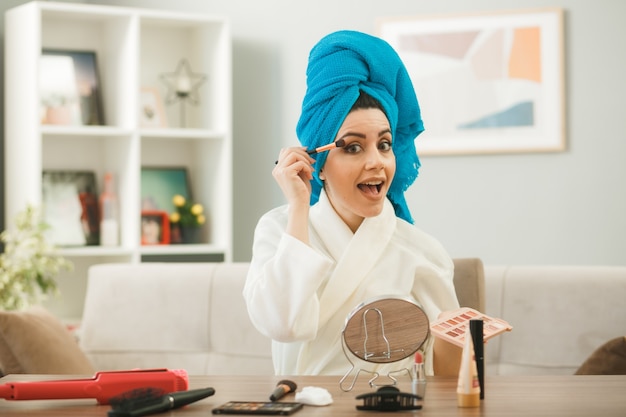 Aufgeregtes junges Mädchen, das Lidschatten mit Make-up-Pinsel aufträgt, das am Tisch mit Make-up-Tools im Wohnzimmer sitzt