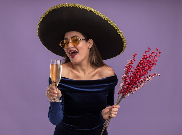 Aufgeregtes junges schönes Mädchen, das blaues Kleid und Brille mit Sombrero hält Ebereschenzweig mit Glas Champagner lokalisiert auf lila Hintergrund trägt