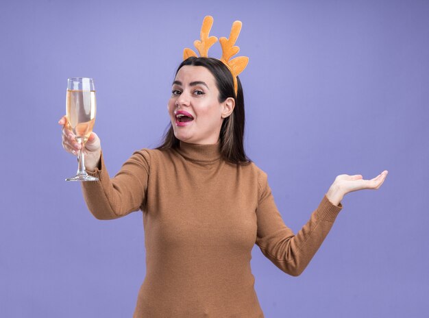Aufgeregtes junges schönes Mädchen, das braunen Pullover mit Weihnachtshaarbügel hält, der Glas der Champagnerverbreitungshand lokalisiert auf blauem Hintergrund hält