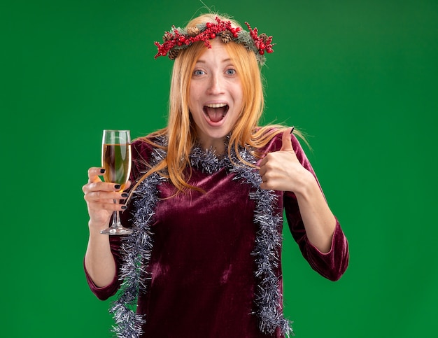 Aufgeregtes junges schönes Mädchen, das rotes Kleid mit Kranz und Girlande am Hals hält Glas des Champagners zeigt Daumen oben isoliert auf grüner Wand
