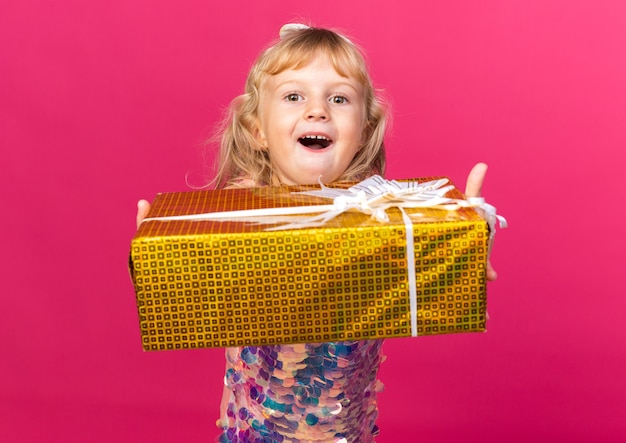 Aufgeregtes kleines blondes Mädchen, das Geschenkbox isoliert auf rosa Wand mit Kopienraum hält