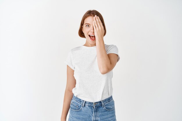 Aufgeregtes Mädchen bedeckt die Hälfte von Gesicht und Auge, keucht erstaunt und sieht etwas Tolles an, steht in T-Shirt und Jeans auf weißer Wand