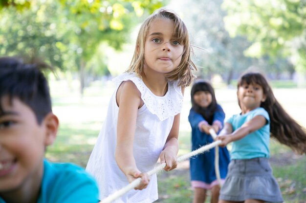 Aufgeregtes Mädchen, das Aktivitäten im Freien mit Klassenkameraden genießt und Tauziehen mit Freunden spielt. Gruppe von Kindern, die Spaß im Park haben. Kindheitskonzept