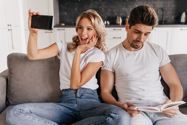 Aufgeregtes Mädchen, das Selfie im Wohnzimmer macht. Kaukasischer Mann, der Buch liest, während er auf Couch sitzt.