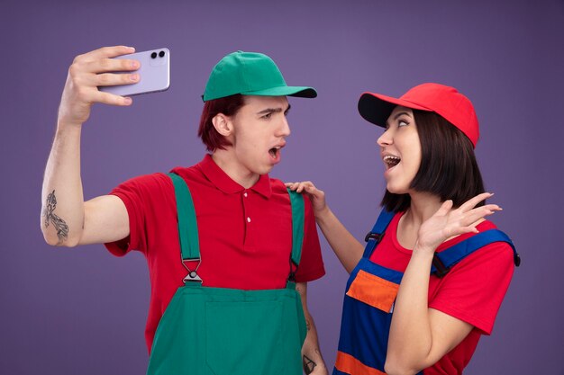 Aufgeregtes Mädchen des jungen Paares ärgerte Kerl in Bauarbeiteruniform und Mütze, die selfie zusammen nehmen und einander Mädchen betrachten, das Hand auf der Schulter des Kerls hält und leere Hand zeigt