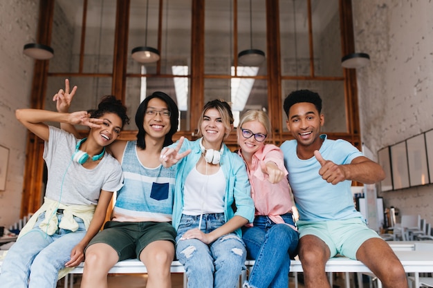 Aufgeregtes Mädchen im blauen Hemd, das Friedenszeichen zeigt, das die Gesellschaft des Freundes an einem guten Tag genießt. Innenporträt von fröhlichen internationalen Studenten, die für Foto herumalbern und lachen ..