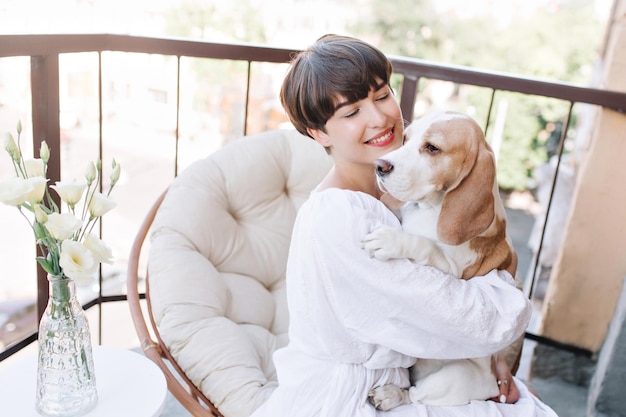 Aufgeregtes Mädchen mit sniny schwarzem Haar, das Beagle-Hund hält, das Vase mit weißen Blumen betrachtet. Porträt einer lächelnden Frau, die im runden Stuhl sitzt, während der Welpe einen guten Tag genießt.