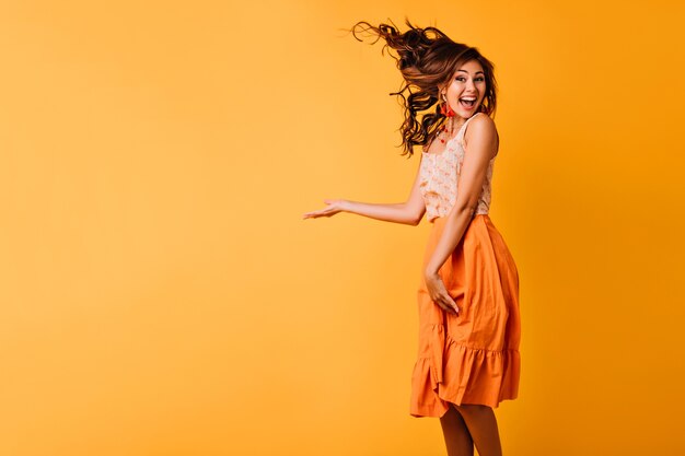 Aufgeregtes Mädchen mit welligem Ingwerhaar, das auf Gelb springt. Studioporträt der glückseligen jungen Frau in der orange Kleidung, die mit Lächeln tanzt.