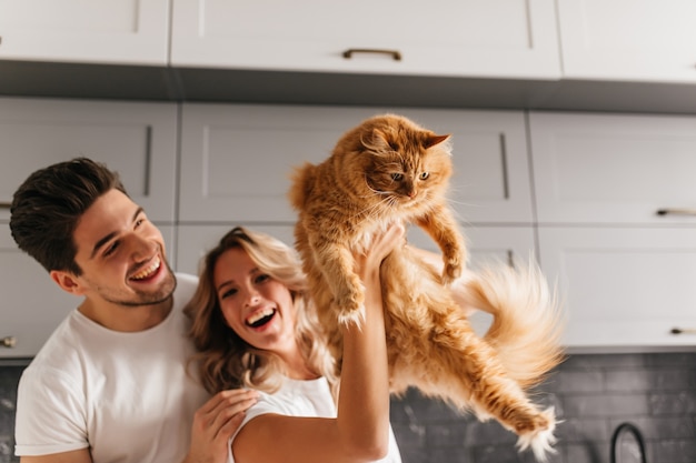 Aufgeregtes Paar, das mit flauschiger Katze aufwirft. Innenporträt der lächelnden entzückenden Frau, die ihr Haustier in der Küche hält.