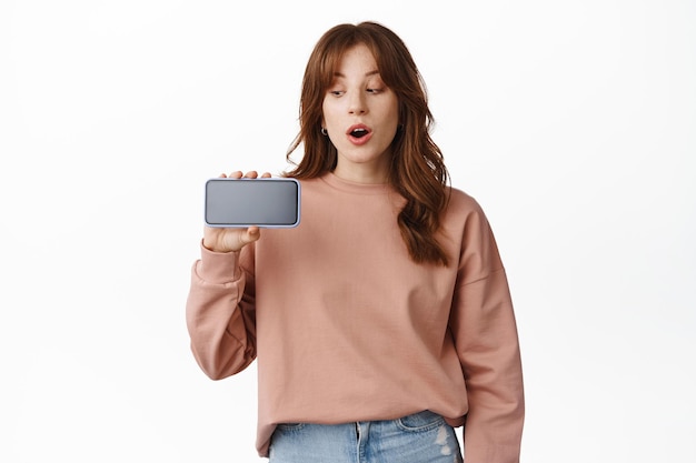 Aufgeregtes rothaariges Mädchen keucht, starrt horizontal beeindruckt auf den Smartphone-Bildschirm, sagt wow, zeigt das Handy und steht vor weißem Hintergrund