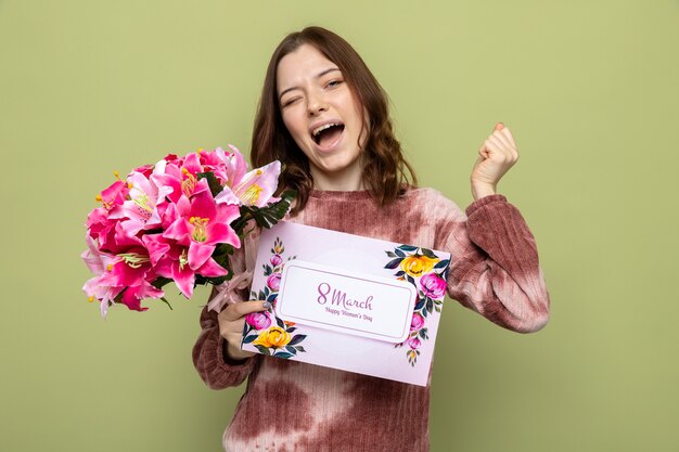 Aufgeregtes schönes junges Mädchen am glücklichen Frauentag, der Blumenstrauß mit Grußkarte hält