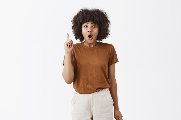 Aufgeregtes und begeistertes kreatives künstlerisches dunkelhäutiges Mädchen mit Afro-Frisur, das vorschlägt, den Zeigefinger in der Eureka-Pose zu heben, die Lippen zu falten und nach Luft zu schnappen, um ihre Idee oder ihren Plan für das Team zu erzählen