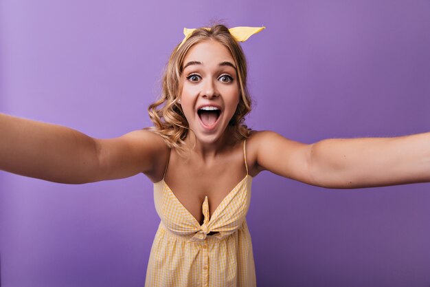 Aufgeregtes weißes weibliches Modell mit gelbem Band, das Selfie macht. Sorglose blonde Frau, die sich auf lila fotografiert.