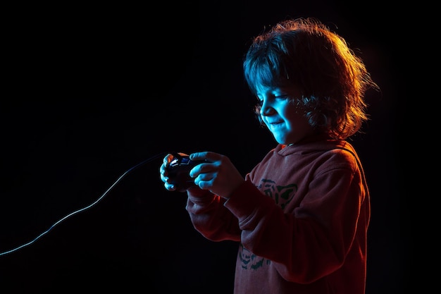 Aufmerksamkeit beim Spielen von Videospielen. Porträt des kaukasischen Jungen auf dunklem Studiohintergrund im Neonlicht. Schönes lockiges Modell.