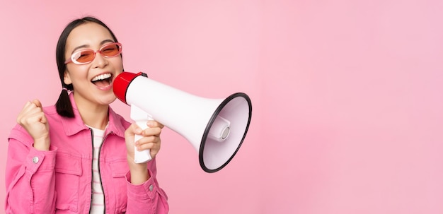 Kostenloses Foto aufmerksamkeitsankündigungskonzept begeistertes asiatisches mädchen, das in megaphonwerbung schreit, wobei lautsprecher rekrutiert werden, die über rosafarbenem hintergrund stehen