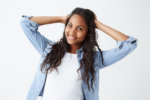 Aufnahme einer gut aussehenden attraktiven jungen afroamerikanischen Frau in Jeanshemd und weißem T-Shirt, die mit ihren langen gewellten Haaren spielt und mit ihren perfekten weißen Zähnen breit lächelt.
