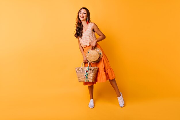 Aufnahme einer inspirierten Frau in voller Länge mit Sommeraccessoires. Glückliches weibliches Ingwermodell im orangefarbenen Rock, der Hut und Tasche hält.