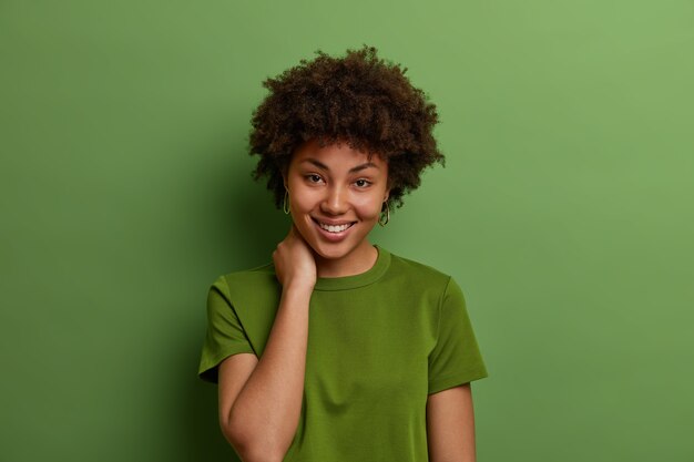 Aufnahme einer schönen Frau mit perfekter Haut und zahnigem Lächeln, berührt den Hals, ist gut gelaunt, hat ein ungezwungenes Gespräch, trägt ein sommergrünes T-Shirt und posiert im Innenbereich. Positives menschliches Gefühlskonzept