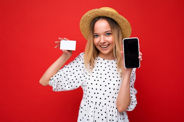 Aufnahme einer schönen glücklich lächelnden blonden frau in sommerkleidung, die smartphone mit leerem bildschirm zeigt