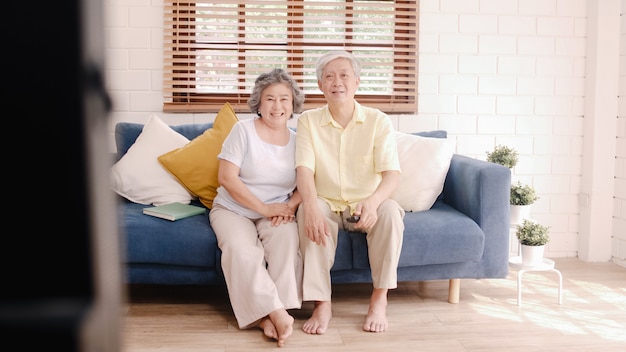 Aufpassendes Fernsehen der asiatischen älteren Paare im Wohnzimmer zu Hause, süße Paare genießen Liebesmoment beim auf dem Sofa liegen, wenn Sie zu Hause entspannt werden.