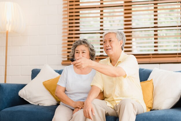 Aufpassendes Fernsehen der asiatischen älteren Paare im Wohnzimmer zu Hause, süße Paare genießen Liebesmoment beim auf dem Sofa liegen, wenn Sie zu Hause entspannt werden.