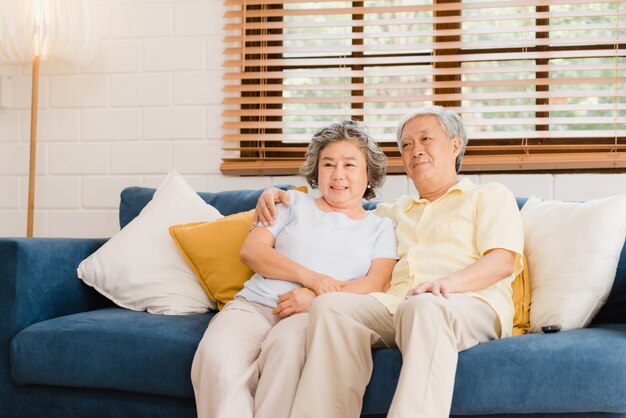 Aufpassendes Fernsehen der asiatischen älteren Paare im Wohnzimmer zu Hause, süße Paare genießen Liebesmoment beim auf dem Sofa liegen, wenn Sie zu Hause entspannt werden.