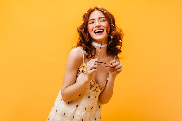 Aufrichtige Frau mit Blumen in roten Locken lacht mit geschlossenen Augen. Frau im gelben Sommerkleid hält Brille auf orange Hintergrund.