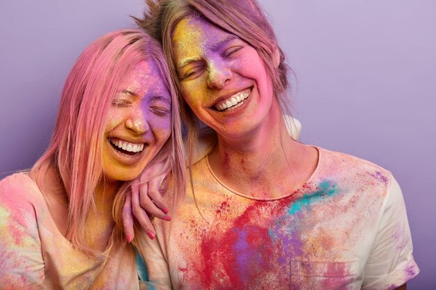 Aufrichtiges Gefühls- und Gefühlskonzept. Lustige zwei Freundinnen lehnen sich aneinander, haben ein breites Lächeln, farbige schmutzige Gesichter, bespritzte Kleidung, nehmen am Holi-Festival teil
