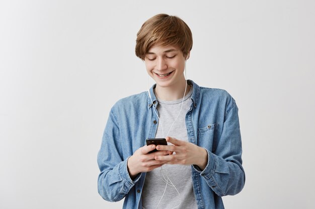 Сaukasischer Kerl mit hellem Haar, das gegen grauen Hintergrund steht, der Smartphone hält, das Musik unter Verwendung der Internetverbindung herunterlädt, die erfreut, aufgeregt, lächelnd schaut, während auf Bildschirm des Handys schaut