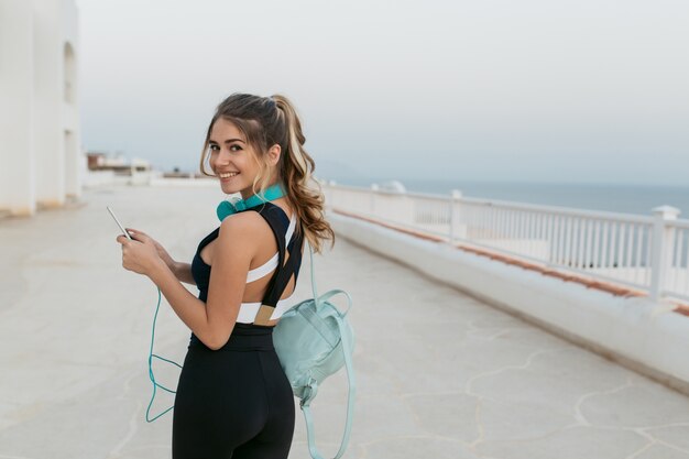 Ausdruck wahrer positiver Gefühle der freudigen jungen Frau in der Sportbekleidung, die am Meer geht. Outwork, sportlicher Lebensstil, modisches Model, Spaß haben, lächeln, telefonieren