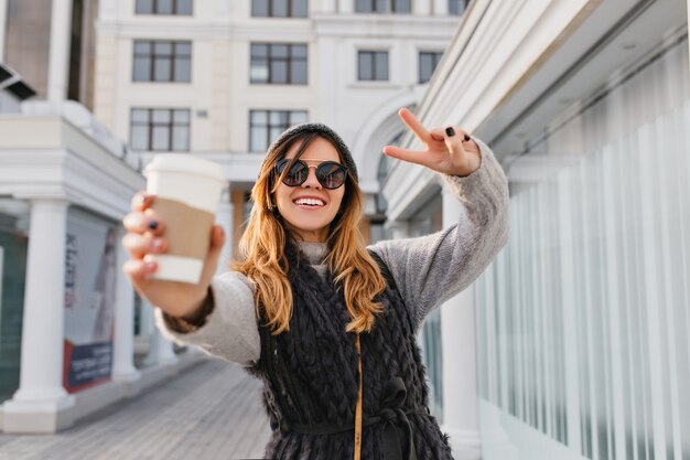 Ausdrücken von hellen positiven Emotionen der modischen Stadtfrau, die Kaffee streckt, um auf sonniger Straße zu gehen. Schöne lächelnde Frau in der modernen Sonnenbrille, Strickmütze, die Spaß im Freien hat.