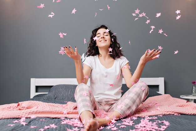 Ausdrücken wahrer positiver Gefühle der jungen freudigen Frau im Pyjama mit geschnittenem lockigem Haar, das Spaß daran hat, rosa Lametta auf Bett in moderner Wohnung zu fallen. Gemütlichkeit zu Hause, lächelnd mit geschlossenen Augen