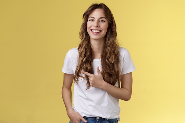 Ausgehende charmante glückliche hübsche Frau lockige lange Frisur, die mit dem Zeigefinger nach links zeigt, zeigt Werbung an, die sorglos lacht, zufrieden, die Handtasche beiläufig freundliche Pose gelber Hintergrund hält.