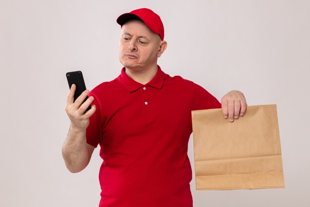 Auslieferungsmann in der roten Uniform und in der Kappe, die Papierpaket hält, das Bildschirm seines Handys mit ernstem Gesicht betrachtet, das über weißem Hintergrund steht