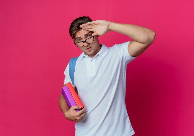 Aussehender junger gutaussehender männlicher Student mit Rückentasche und Brille, der Bücher hält und Grußgeste zeigt