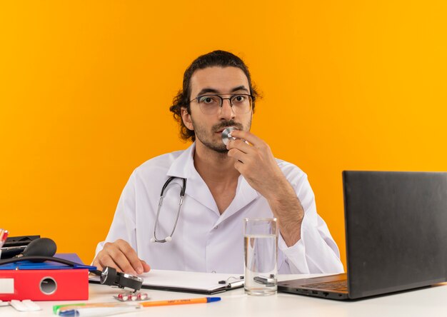 Aussehender junger männlicher Arzt mit medizinischer Brille, der ein medizinisches Gewand mit Stethoskop trägt, das am Schreibtisch sitzt