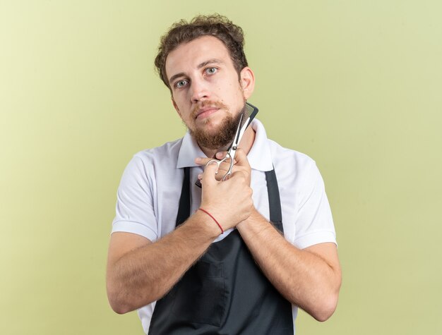 Aussehender junger männlicher Friseur, der einen einheitlichen Kamm mit einer Schere um das Gesicht trägt, isoliert auf olivgrüner Wand