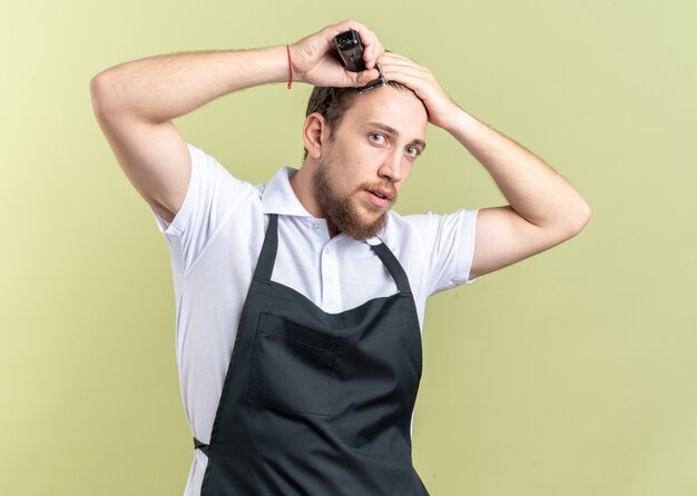 Aussehender junger männlicher Friseur, der einheitliche Haare mit Haarschneidemaschinen trägt, die auf olivgrüner Wand isoliert sind?