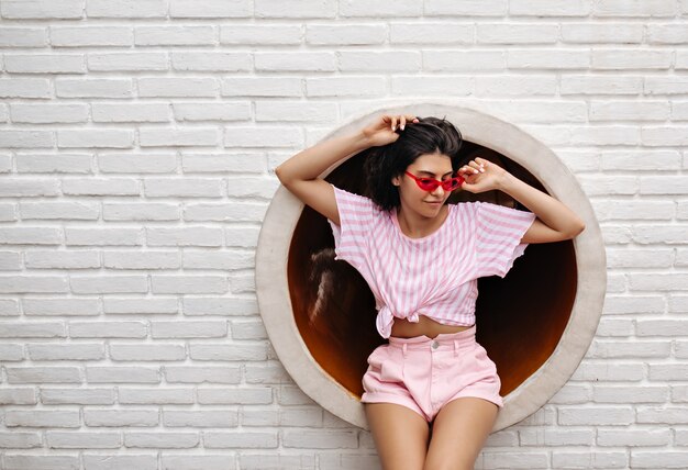 Außenaufnahme der inspirierten Frau in der Sonnenbrille. Brünette Frau in der Sommerkleidung, die auf städtischem Hintergrund aufwirft.