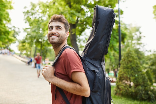 Außenaufnahme des glücklichen gutaussehenden hipster-mannes bewundern schönen grünen park, der mit gitarre geht, musiker, der auf probe geht