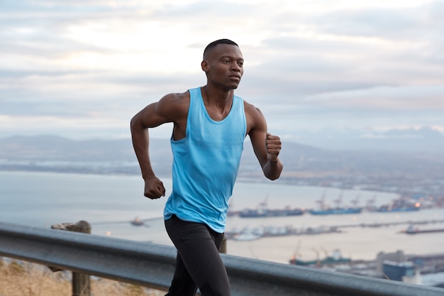 Außenaufnahme des selbstbewussten afroamerikanischen Fitness-Mannes hat Zielerreichung Herausforderung, Ziel ohne Pause zu erreichen, arbeitet aktiv mit den Händen, gekleidet in Sportbekleidung, joggt über schöne Naturansicht