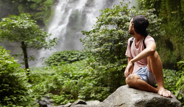 Außenaufnahme des stilvollen jungen kaukasischen Wanderers, der barfuß auf großem Felsen sitzt und über seine Schulter auf herrlichen Wasserfall schaut