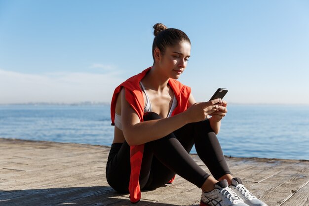 Außenaufnahme einer attraktiven Sportlerin, die eine Pause am Meer macht, die auf einem Holzsteg sitzt und mobi...