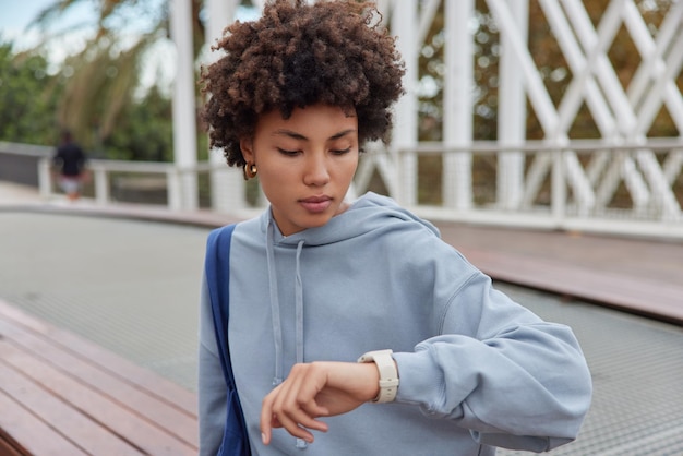 Außenaufnahme einer Frau mit lockigen Haaren, die einen lässigen Hoodie trägt, überprüft die Zeit auf der Smartwatch, trägt eine Tasche, wartet darauf, dass jemand ernsten Gesichtsausdruck hat, wartet darauf, dass der Trainer mit dem Sporttraining beginnt. Menschen und Lebensstil