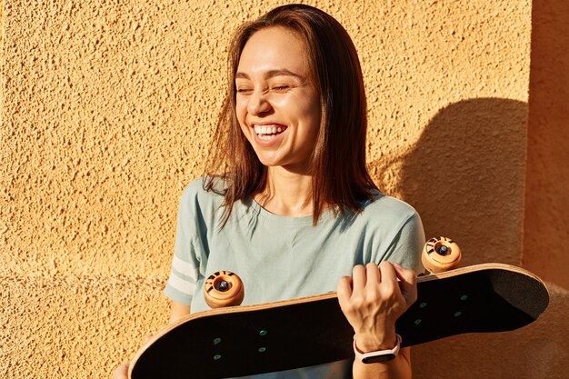 Außenaufnahme einer jungen, dunkelhaarigen, lachenden Frau, die die Augen geschlossen hält, ein T-Shirt trägt, ein Skateboard in den Händen hält und gegen eine gelbe Wand steht.